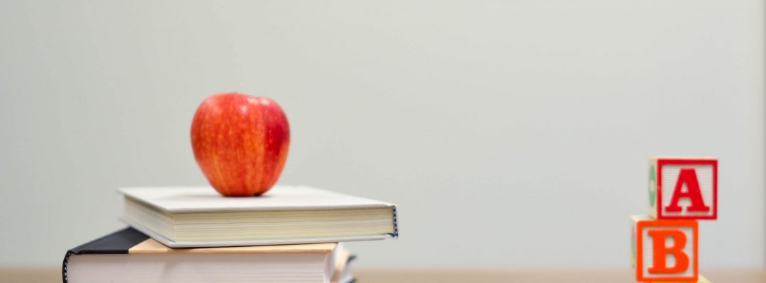 Apple on top of a pile of books next to some coloured pencils and a stack of letter blocks symbolising entry into a classroom